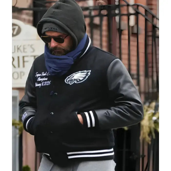 Person wearing a black varsity jacket with a logo, blue scarf, and dark sunglasses outdoors.
