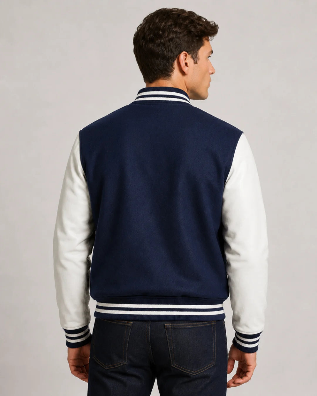 Back view of mens navy blue varsity jacket with blank wool body, white leather sleeves, and striped ribbed hem on male model